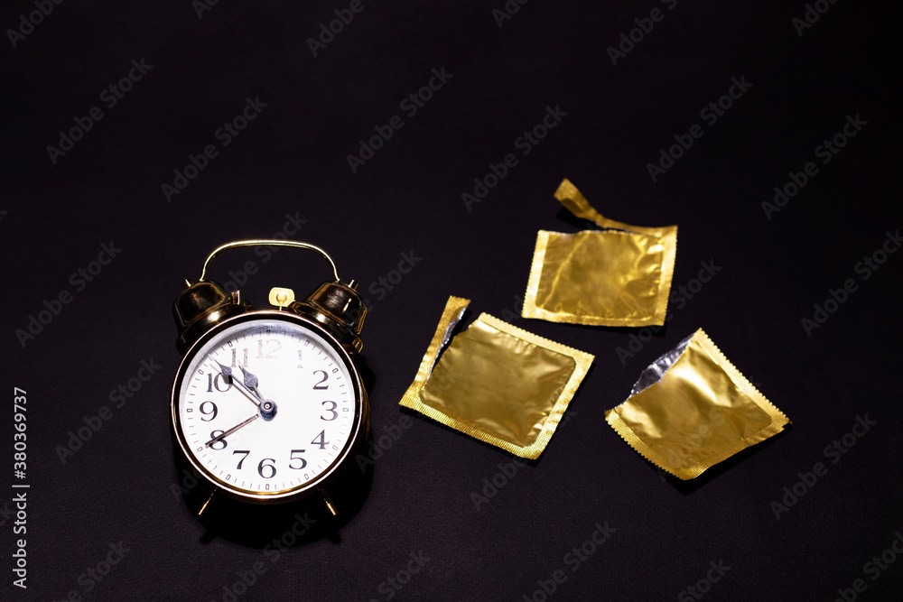 A gold watch is located on a black background next to three opened condoms. Special design for the contraception day. There is a space for text. A gold watch is located on a black background next to three opened condoms. Special design for the contraception day. There is a space for text.