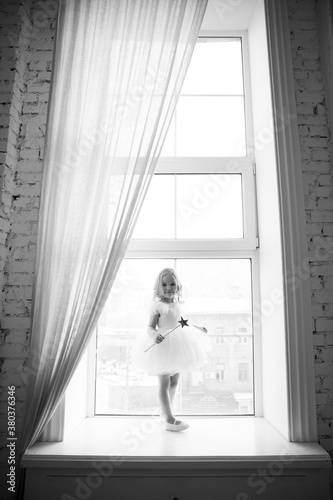 a little girl in a smart, fluffy white dress with a magic wand in her hands near the window and waits for Santa Claus or the tooth fairy. children's fairy tales and dreams. new year holiday.