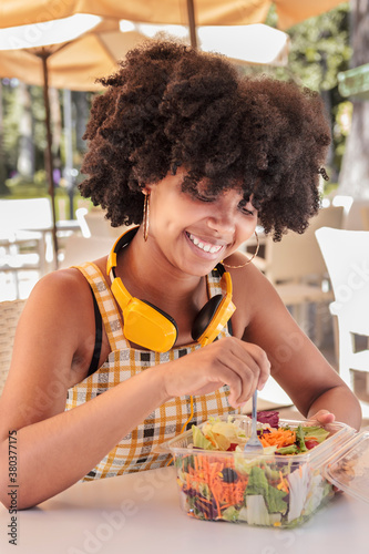 Young African American woman with curly hair and headphones smiles as she eats salad in a plastic to-go container on the terrace of an outdoor cafe on a sunny day. Diet and food for healthy living.