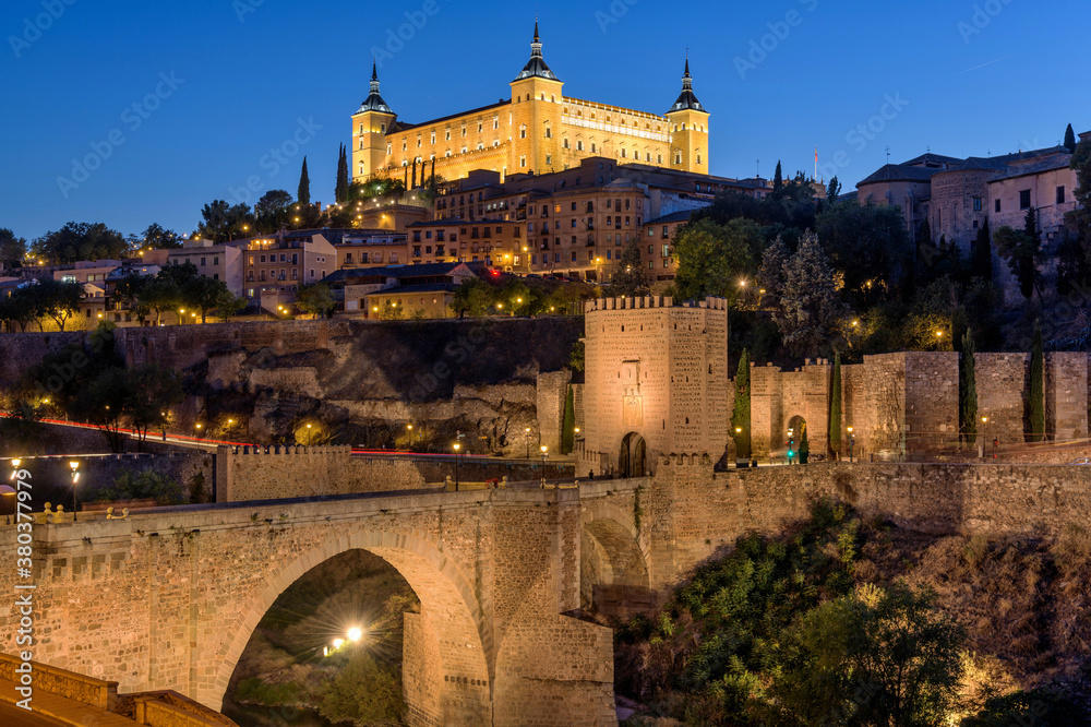 Toledo at Dusk - A panoramic dusk view of the historic city Toledo at Puente de Alcántara - an ancient Roman arch bridge located in front of eastern city gate Puerta de Alcántara. Toledo, Spain.
