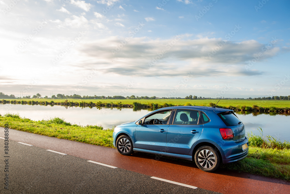 A blue parked car on the side of the road near the ringvaart canal in the North-Holland village of Abbenes on a cloudy day in the Netherlands.