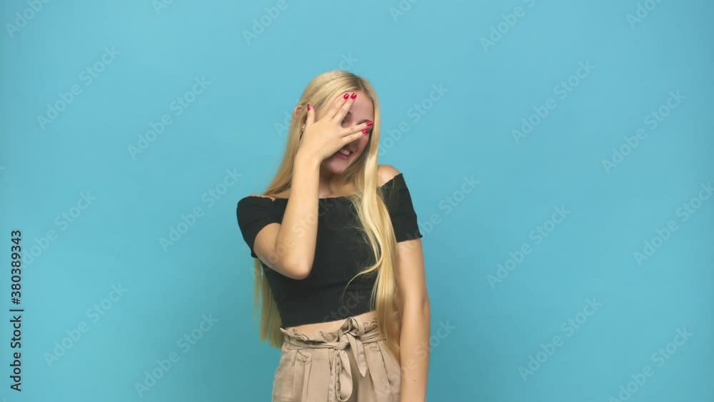 Young caucasian blonde woman blink at the camera through fingers, embarrassed covering face