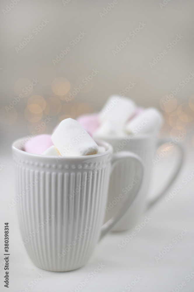 Hot winter drink with cocoa and marshmallows in mugs on wooden white background with Christmas garland. Top view.
