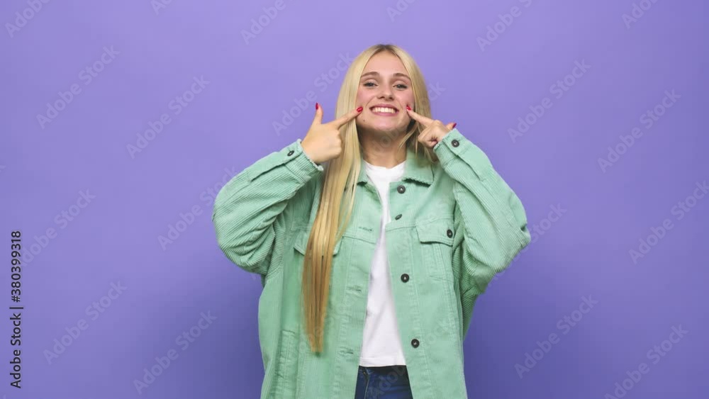 Young caucasian blonde woman smiles, pointing fingers at mouth Young caucasian blonde woman smiles, pointing fingers at mouth