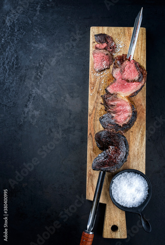 Barbecue dry aged wagyu Brazilian picanha from the sirloin cap of rump beef sliced and offered as top view on a skewer on a wooden design board on rustic black background with copy space photo