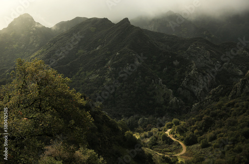 Misty mountain peaks tower over a lone dirt road snaking through the lush valley below, lit by a pool of sunlight . 