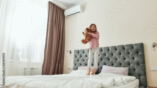 A cute small girl jumping barefoot on a big bed smiling and embracing a brown teddybear