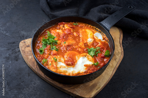 Traditional Israeli national dish shakshouka offered as breakfast with poached eggs in tomato sauce with chili and onions offered as close-up in a design cast iron pan with copy space