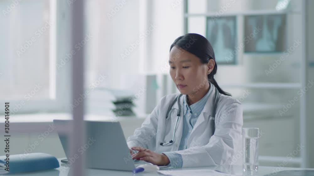 Portrait footage of Asian general practitioner sitting alone in her office working on laptop adding data to online medical card