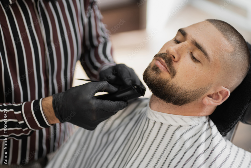 master of men's hairstyles Barber makes a new short stylish beard haircut with a special professional machine, a client is a young man with a dark-haired beard of Caucasian appearance master of men's hairstyles Barber makes a new short stylish beard haircut with a special professional machine, a client is a young man with a dark-haired beard of Caucasian appearance