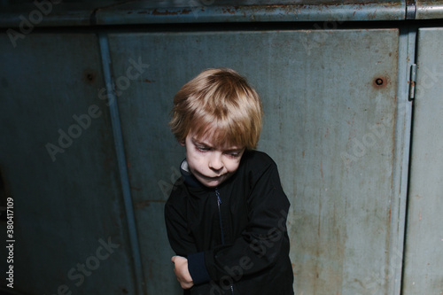 A thoughtful child looking down wearing a black jacket against grey-blue factory walls.