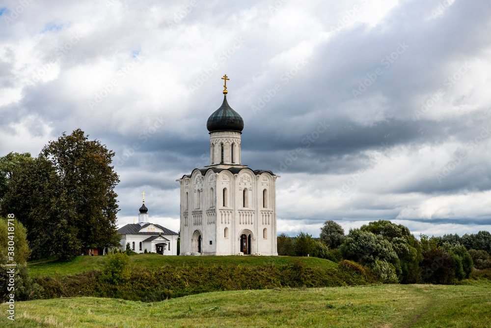 snow-white stone old church against the background of a green meadow and lake before a summer thunderstorm