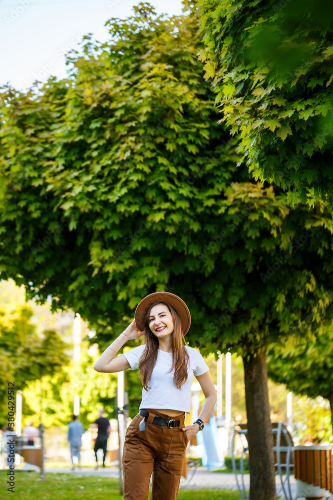 Fototapeta premium Young happy woman in a hat walks along an alley in a park. A girl of European appearance with a smile on her face on a bright sunny summer day