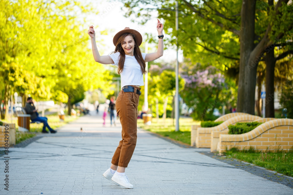 Young happy woman in a hat walks along an alley in a park. A girl of European appearance with a smile on her face on a bright sunny summer day