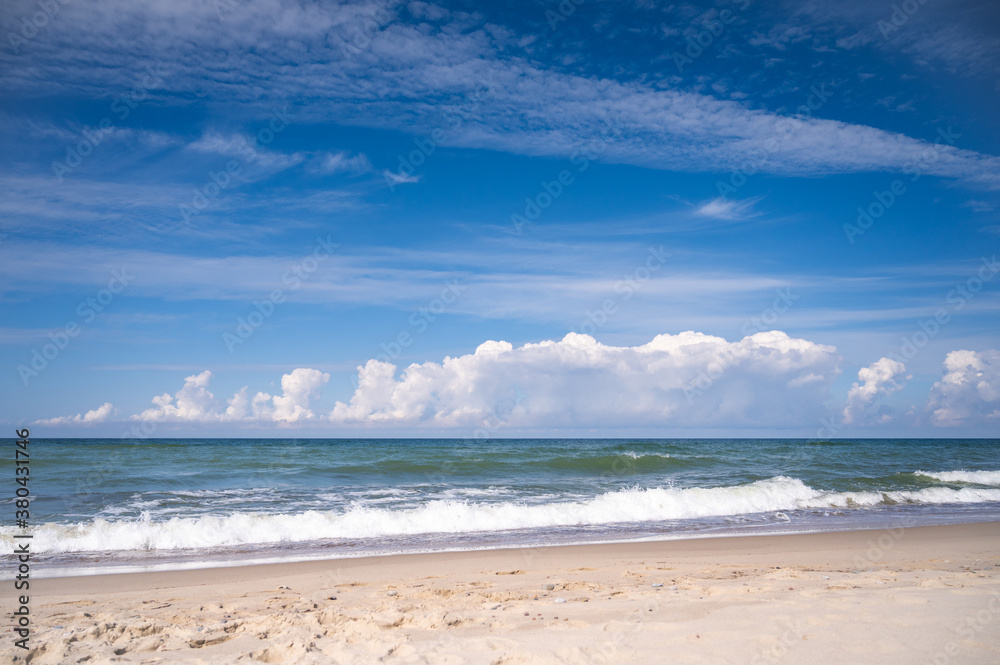 Panorama of a deserted sandy beach on which the sea foamy waves roll. An endless blue sky with white clouds on it.