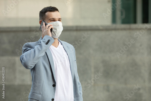 European young man in medical mask on his face calling by phone. Man in protective mask is protected from virus outdoor in street. Covid-19.