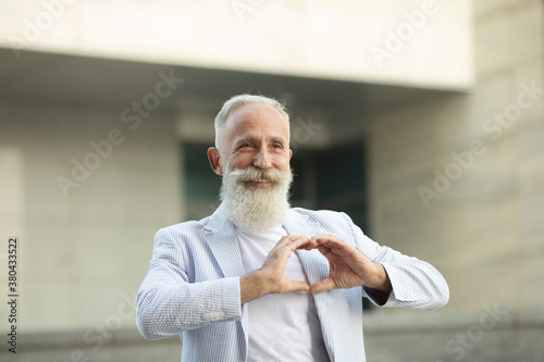 Lovely good-looking caucasian bearded senior man and making heart gesture, expressing his feelings and tenderness while standing against street background. I am in love with you.