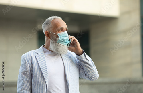Bearded mature man with medical mask using mobile phone outdoor. Covid-19.