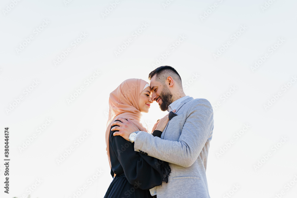 Muslim love story close-up with sun light. Mixed couple smiles and hugs at sunset. Woman weared in hijab looks to her man. Advert for on-line dating agency