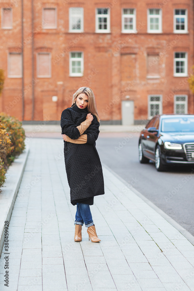 Outdoors lifestyle fashion portrait of stunning blonde young woman walking on the street. Wrapping up in a coat from the cold. Wearing stylish black coat and ankle boots. Cold weather. Feeling cold