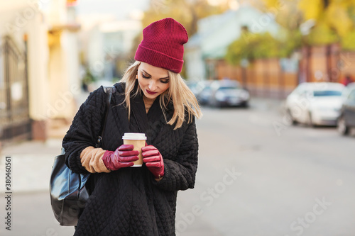 Outdoors lifestyle fashion portrait of stunning blonde girl. Smiling, drinking coffee and walking on the city street. Going shopping. Trendsetter. Wearing stylish black oversized coat and burgundy hat
