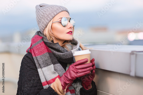 Outdoors lifestyle fashion portrait of stunning blonde girl. Sitting in an outdoor cafe on the roof. Drinking coffee and enjoying life. Wearing stylish black oversized coat, scarf, sunglasses and hat © Olesya Kuprina