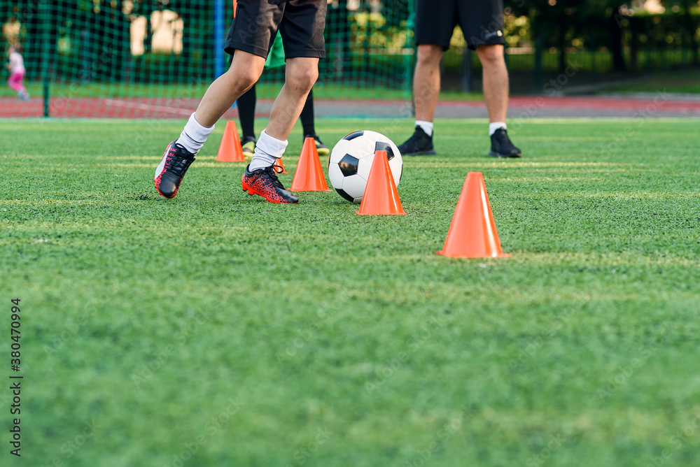 Obraz premium Close up boy in sportswear trains football on soccer field and learns to circle the ball between training orange cones.