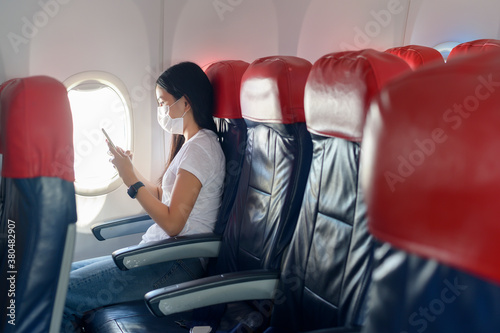 Traveling woman wearing protective mask onboard in the aircraft using smartphone, travel under Covid-19 pandemic, safety travels, social distancing protocol, New normal travel concept