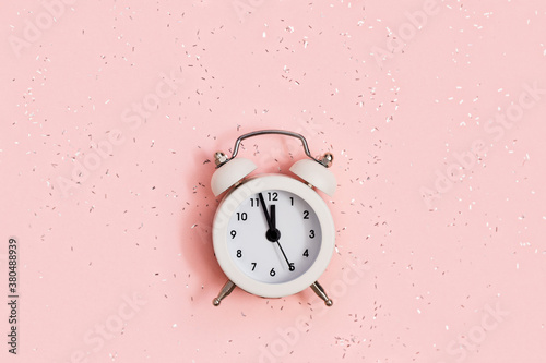 White alarm clock shows about twelve oсlock on pink paper background covered with sequins with copy space. New year Christmas concept. Flat lay. Top view.
