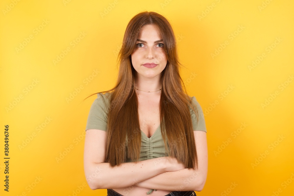 Obraz premium Young caucasian woman with long hair wearing green tshirt standing over isolated yellow background serious face with crossed arms looking at the camera. Positive person.