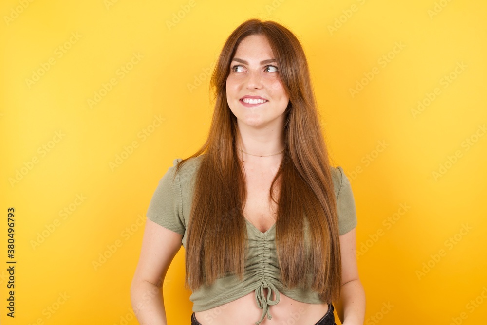 Young caucasian woman with long hair wearing green tshirt standing over isolated yellow background bitting lip and looking up to empty space, Young caucasian woman with long hair wearing green tshirt standing over isolated yellow background bitting lip and looking up to empty space,