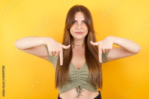 Young caucasian woman with long hair wearing green tshirt standing over isolated yellow background pointing with two fingers at copy space down. Place for advertising