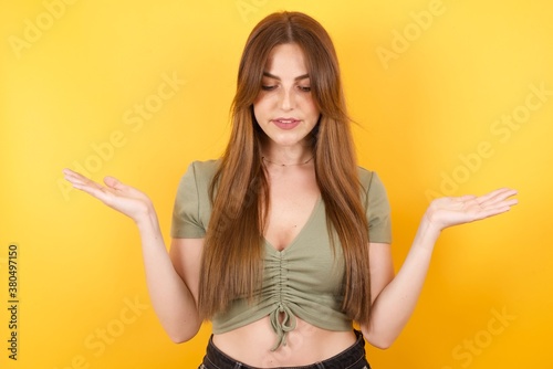 Hesitant Young caucasian woman with long hair wearing green tshirt standing over isolated yellow background shrugs shoulders, looks uncertain and confused. Have no answer © Roquillo