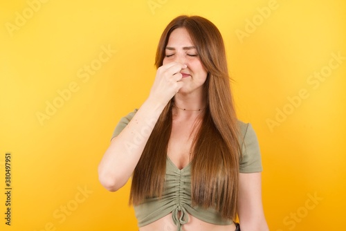 Young caucasian woman with long hair wearing green tshirt standing over isolated yellow wall smelling something stinky and disgusting, intolerable smell, holding breath with fingers on nose. Bad smell