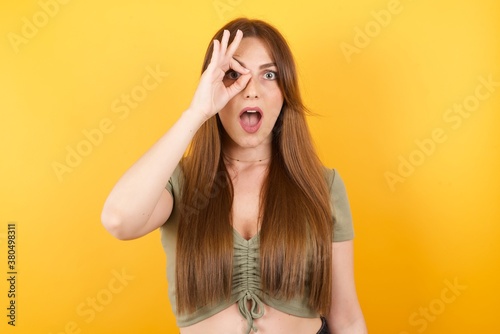 Young caucasian woman with long hair wearing green tshirt standing over isolated yellow background doing ok gesture shocked with surprised face, eye looking through fingers. Unbelieving expression.