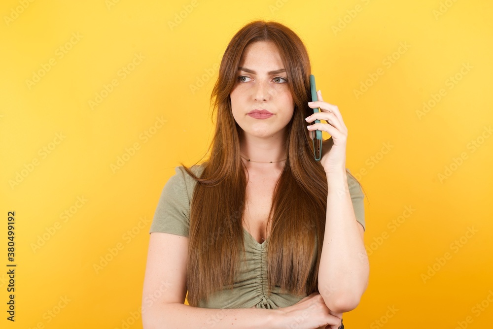 Fototapeta premium Sad brunette caucasian woman in red shirt talking on smartphone on yellow background. Communication concept.