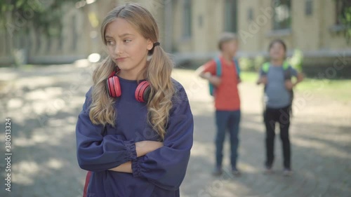 Portrait of sad stressed schoolgirl standing with crossed hands on sunny schoolyard as blurred boys laughing at her at the background. Concept of middle school bullying.