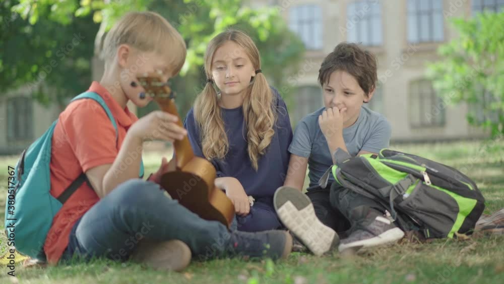 Relaxed Caucasian children sitting on sunny schoolyard and playing ukulele. Portrait of carefree middle school students and friends resting after studying outdoors. Friendship concept. Relaxed Caucasian children sitting on sunny schoolyard and playing ukulele. Portrait of carefree middle school students and friends resting after studying outdoors. Friendship concept.