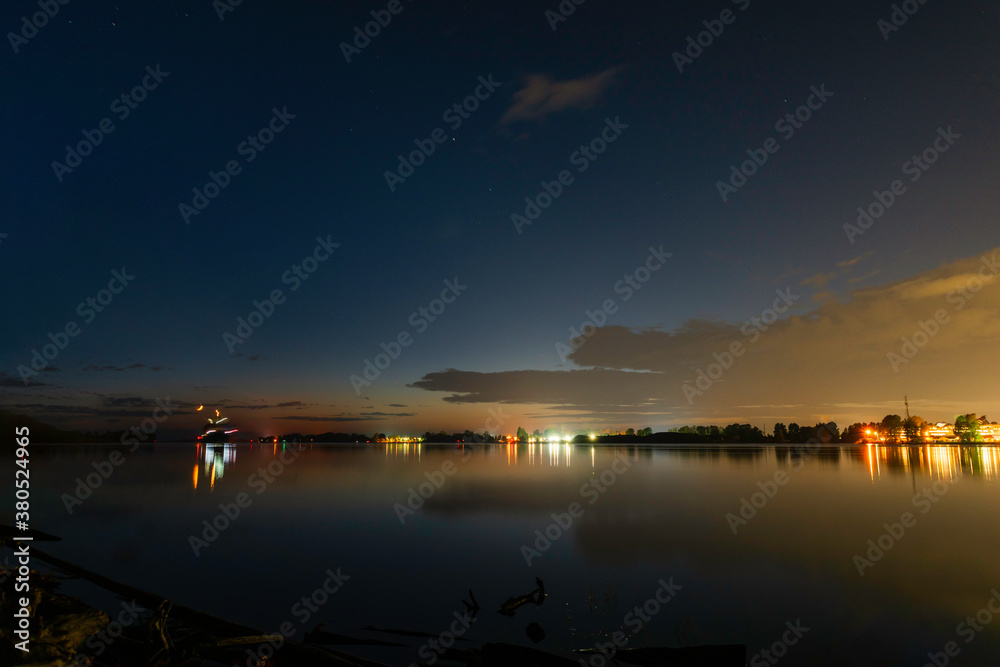 A night with lights and reflection on the river. The landscape of a summer night on the river with lights of lanterns and reflection in the water. Blue sky with clouds and points of stars in the sky
