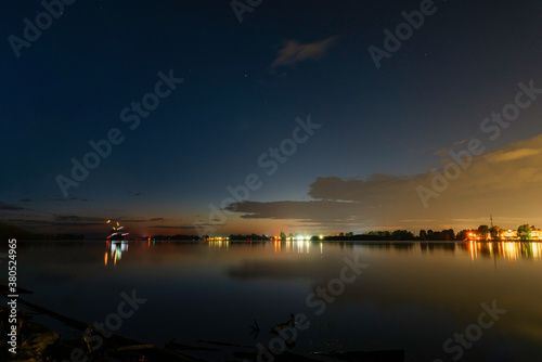 A night with lights and reflection on the river. The landscape of a summer night on the river with lights of lanterns and reflection in the water. Blue sky with clouds and points of stars in the sky