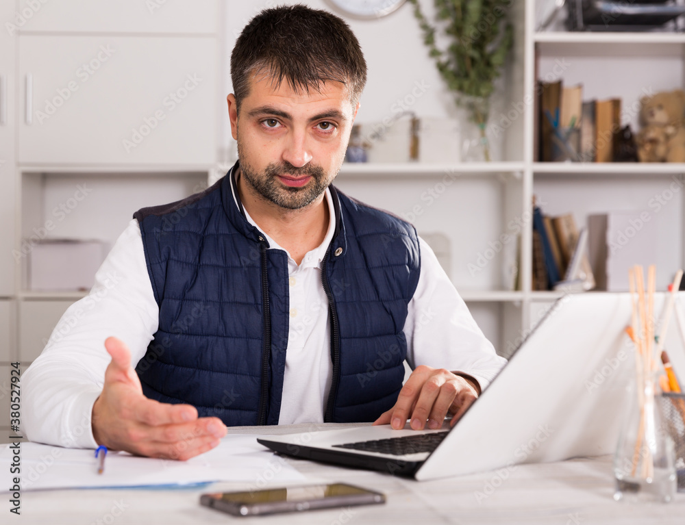 Naklejka premium Portrait of young man with laptop and papers working in office indoors..
