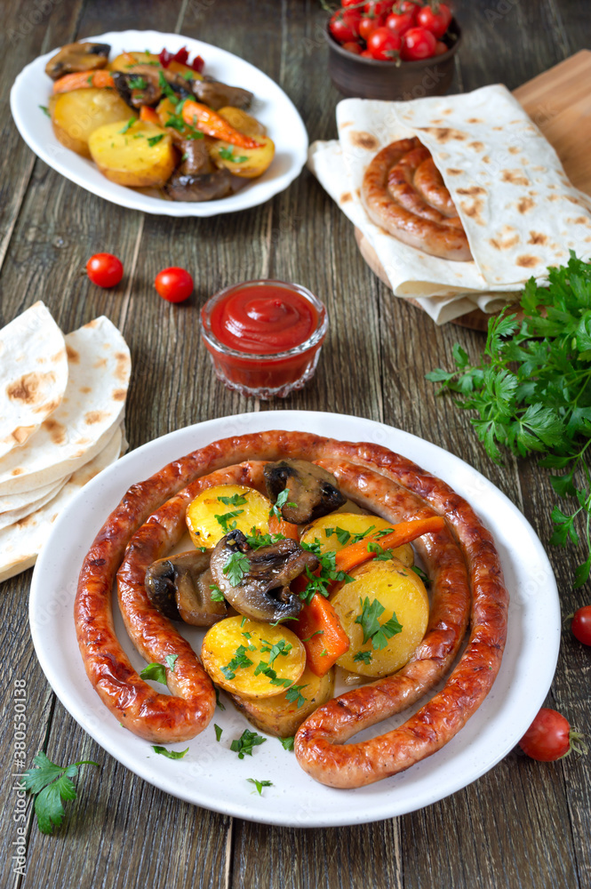 Tasty homemade grilled sausages with baked herb potatoes, mushrooms, cherry tomatoes and ketchup on a wooden background. Oktoberfest snack. Summer picnic dish Tasty homemade grilled sausages with baked herb potatoes, mushrooms, cherry tomatoes and ketchup on a wooden background. Oktoberfest snack. Summer picnic dish