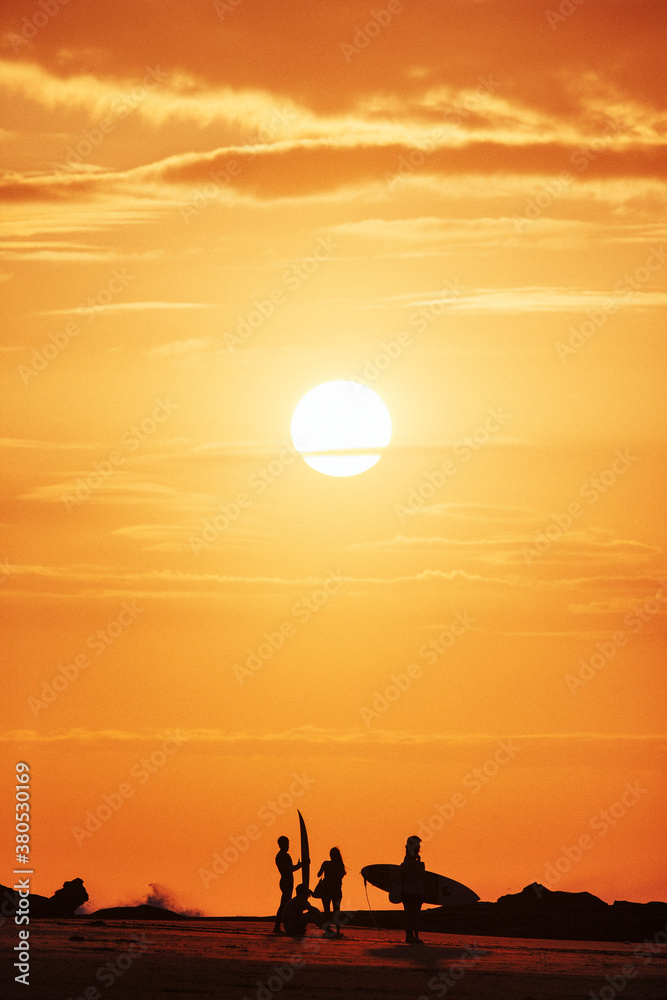 A group of surfers stand silhouetted against a bright orange sunset A group of surfers stand silhouetted against a bright orange sunset