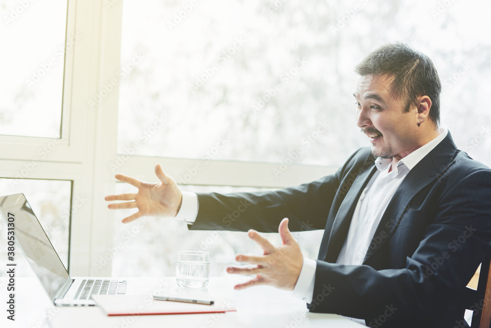 Asian successful businessman sitting at the table with laptop. Panoramic window background. Triumph, happiness. Asian successful businessman sitting at the table with laptop. Panoramic window background. Triumph, happiness.