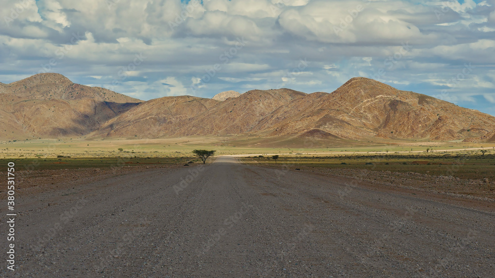 Gravel road with lonely tree covered by cloud shadow and sun shining on round hills and slightly green meadows in the background after rainy season near Solitaire, Namib desert, Namibia, Africa.