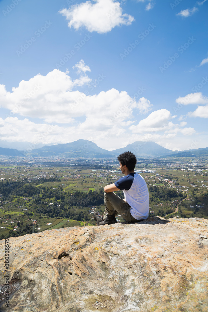 Fototapeta premium Young hispanic man sitting on top of the mountain watching the horizon - man meditating in the heights observing the volcano and mountains in front of him