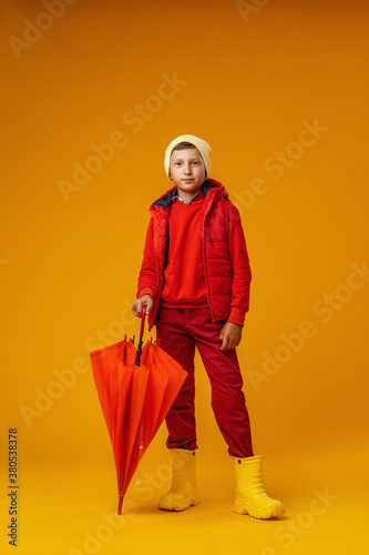 happy emotional boy in autumn clothes with umbrella stands on yellow background