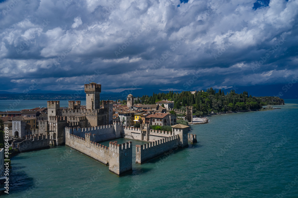 Fototapeta premium Aerial view on Sirmione sul Garda. Italy, Lombardy. Aerial photography with drone. Rocca Scaligera Castle in Sirmione. Cumulus clouds over the island of Sirmione.