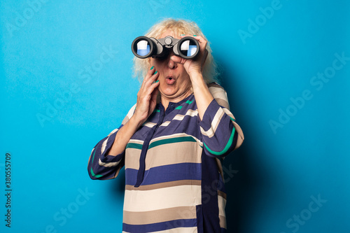 Surprised old woman in a striped dress looks through binoculars on a blue background