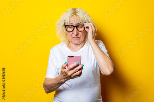 Old woman in white t-shirt with glasses looking at phone on yellow background.